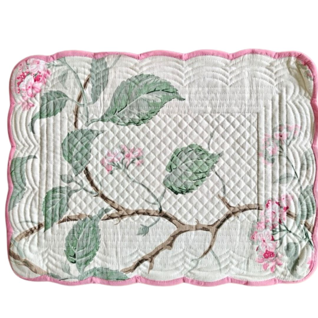 A square quilted placemat with a scalloped pink border, featuring a green leaf and brown branch pattern on a white background with pink floral accents.