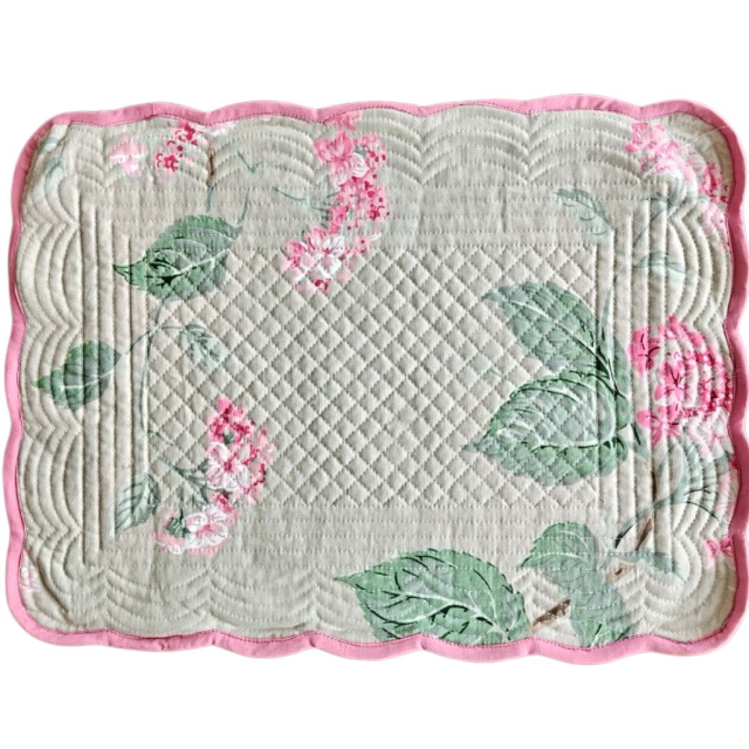 A square quilted fabric placemat with a scalloped pink border, featuring a light gray-green background with a quilted diamond pattern, green leaves, and pink flowers.